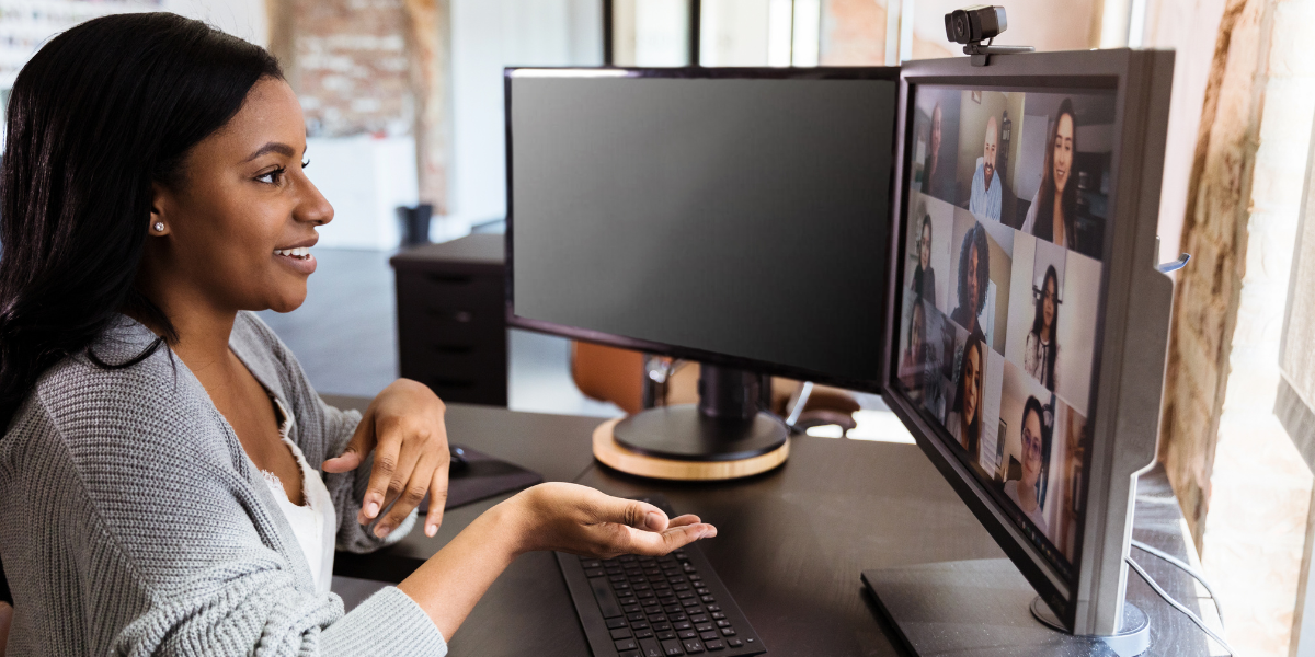 A woman is sat at a desk with two monitors participating in a virtual meeting.