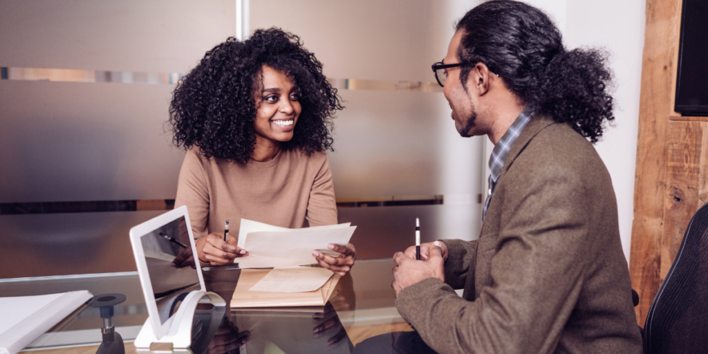 Two people are sat at an office table reviewing documents and conversing.
