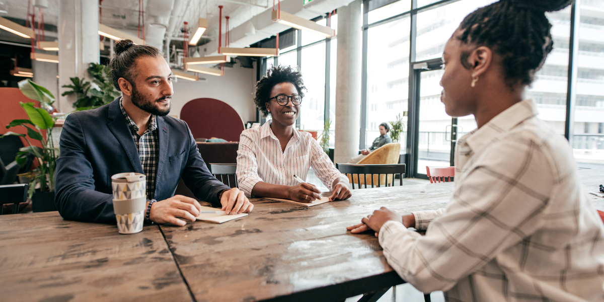 Three people are sat at a boardroom table in front of a wall of windows conversing.