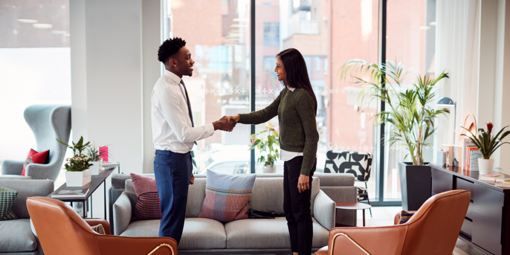 Two people are standing in an office space shaking hands in front of a wall of windows.