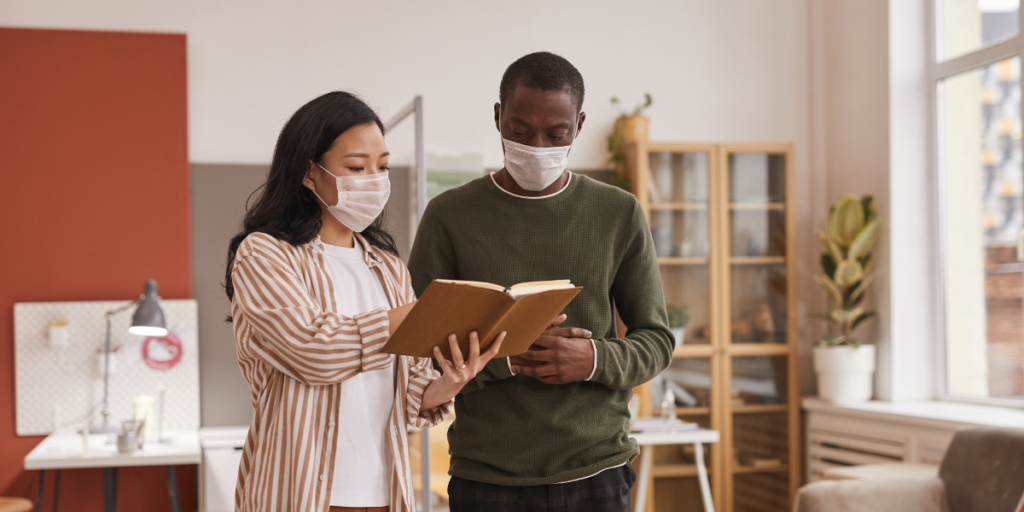 Two people are standing in an office space reviewing notes. They are both wearing medical face masks.