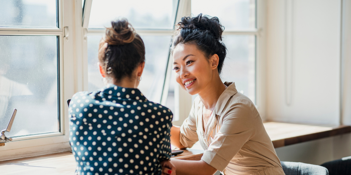 Two women are sat at a table facing one another conversing.