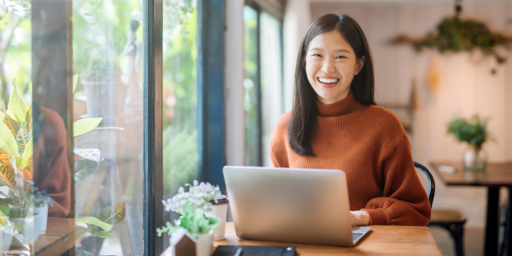 A woman is sat at a desk beside a wall of windows typing on her laptop.
