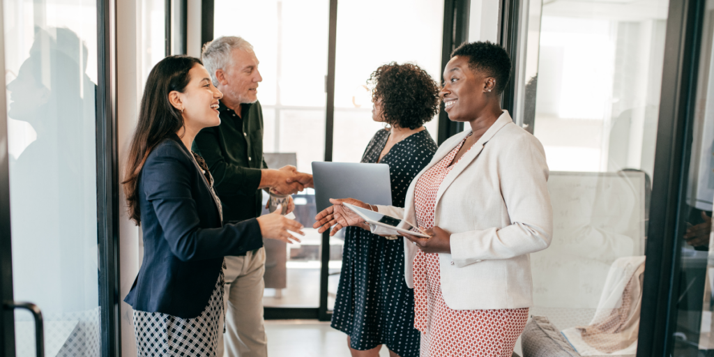 A group of people are standing in an office hallway shaking hands and conversing.