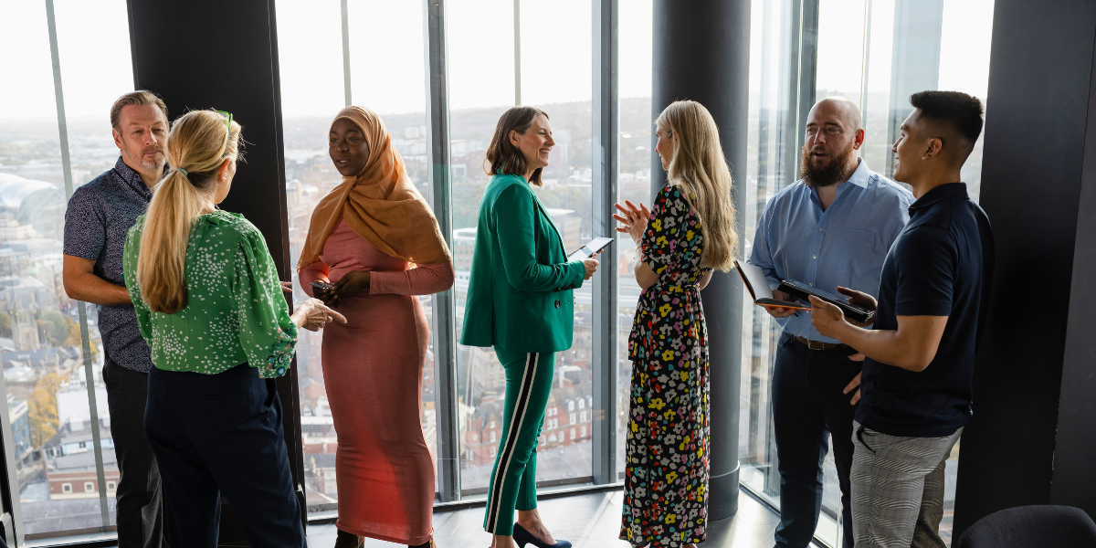 7 people standing in front a wall of windows conversing and 3 of the individuals are holding tablets appearing to interview three of the individuals.