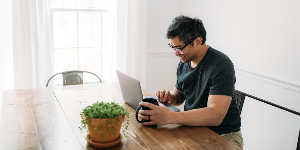A man sitting at a table with a coffee mug typing on his laptop.