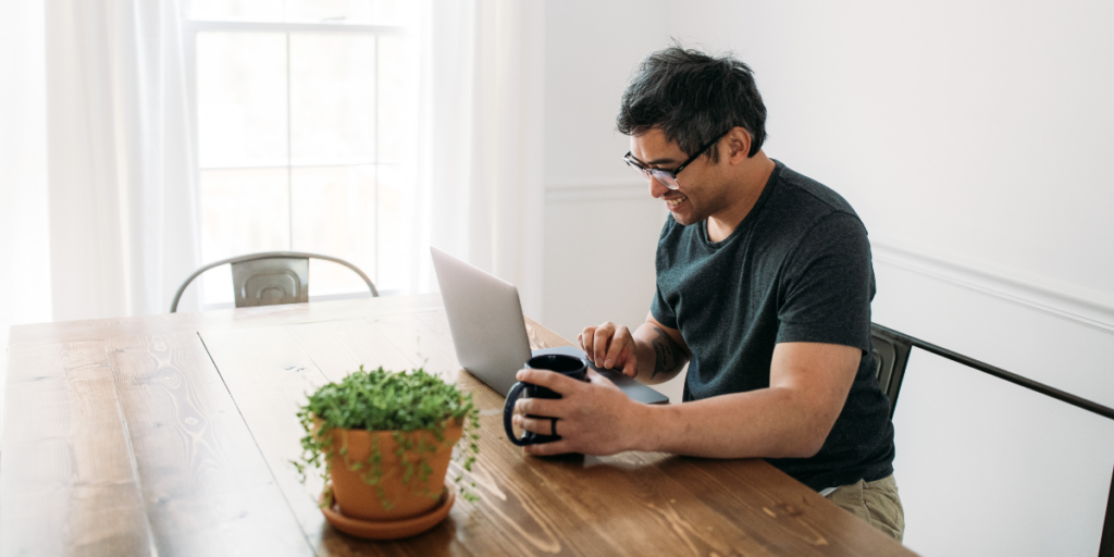 A man sitting at a table with a coffee mug typing on his laptop.