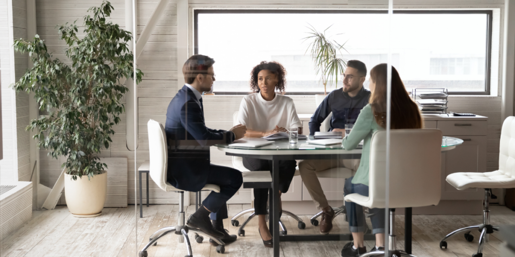 A group of four people sitting in a window-lined office conversing.