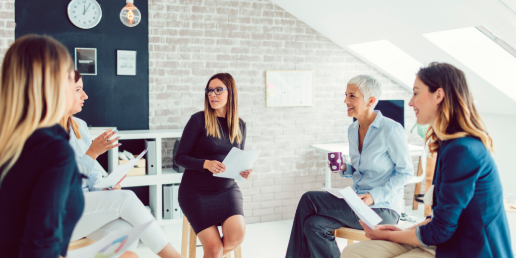 A group of women sitting in an office conversing and holding documents.