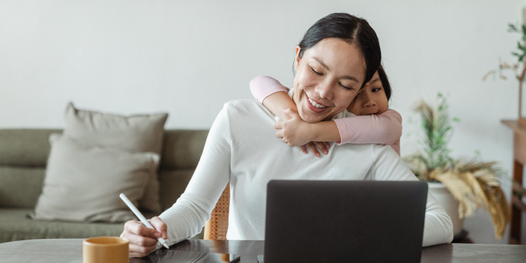 A woman sat at her desk working and her daughter is holding her arms around her.