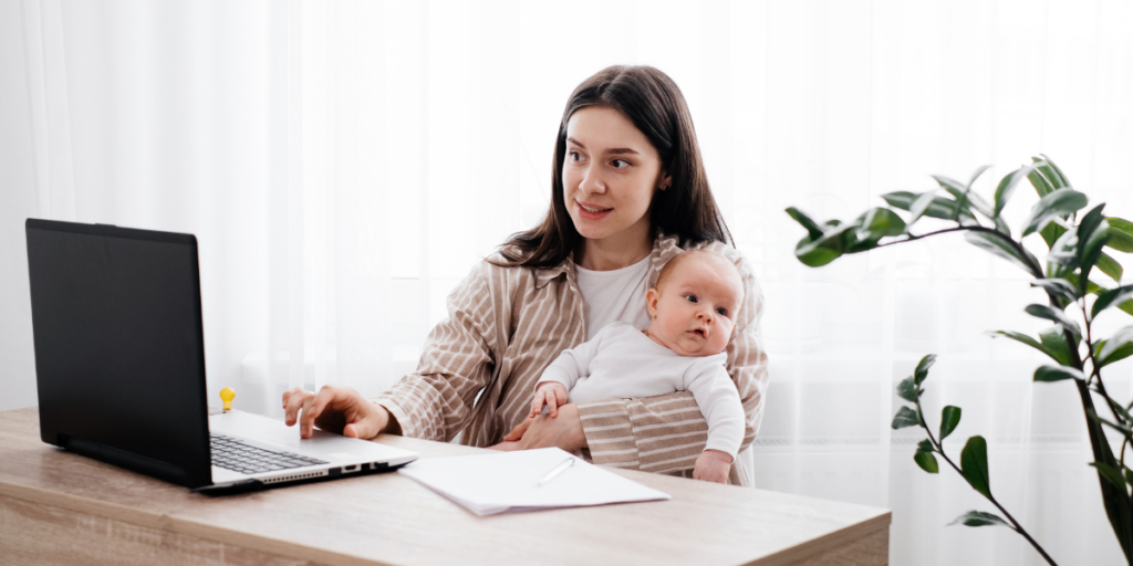 A woman sat at a desk holding a baby and typing.