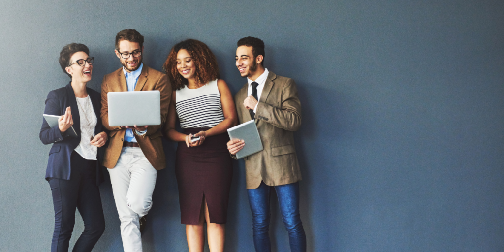 Four people standing against a navy blue wall conversing and reviewing a laptop screen.