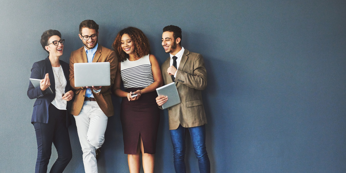 Four people standing against a navy blue wall conversing and reviewing a laptop screen.