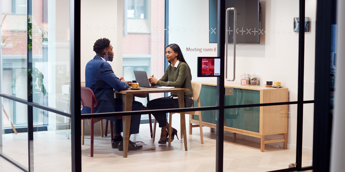 Two people are sat in an office lined with windows conversing.