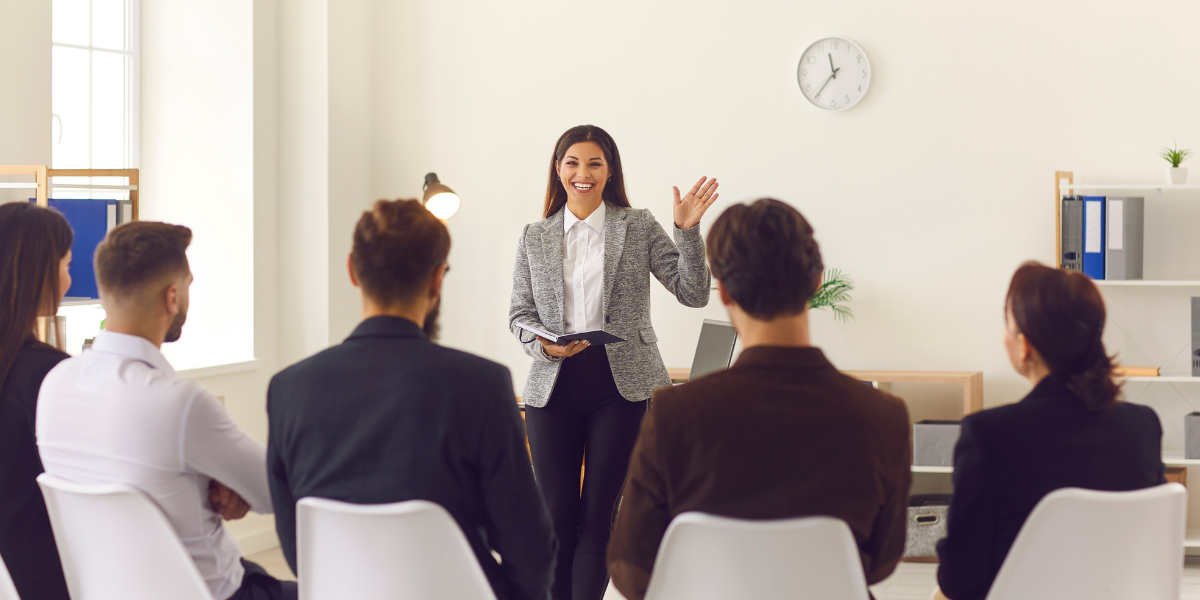 A woman standing at the front of the room holding a notebook and waving to a group of people sitting.