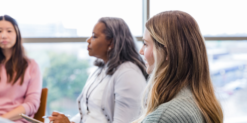 Three people sat in an office near a wall of windows conversing.