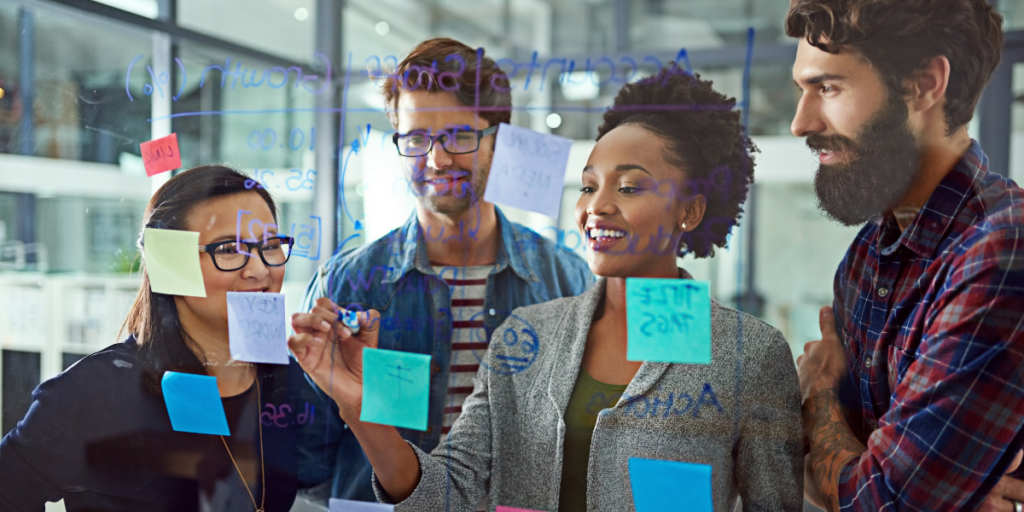 A group of four people standing at a whiteboard watching the one person write notes.