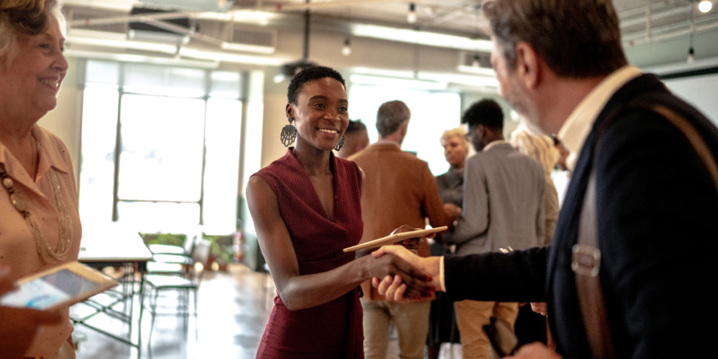A woman is stood in a crowded room shaking the hand of a man facing away from the camera.