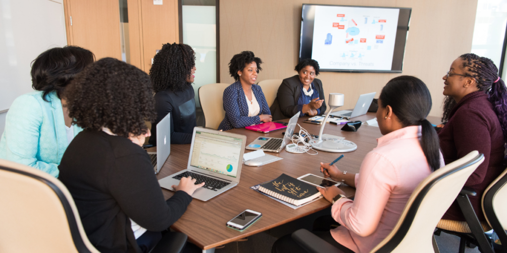 A boardroom of women sitting at the table are conversing.
