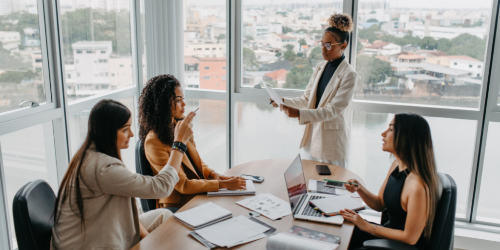 Three women sitting at a boardroom table and one woman is standing presenting to the group.