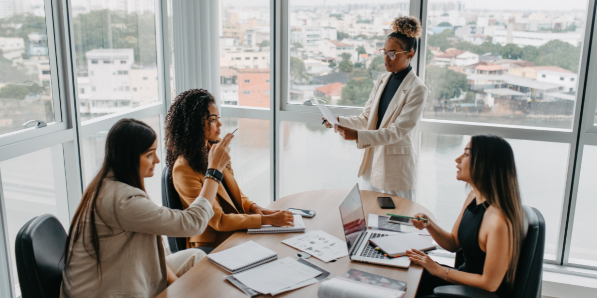 Three women sitting at a boardroom table and one woman is standing presenting to the group.