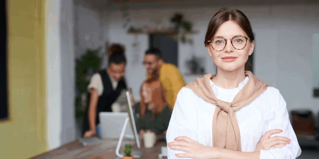 A woman is stood staring at the camera smiling. There is a group of people behind her working.