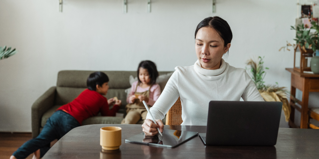 A woman sits at a table working on her tablet while her children are sitting on the couch behind her.