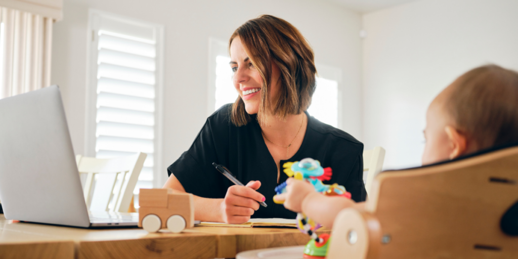 A woman sits at her desk looking at her laptop with her baby next to her.