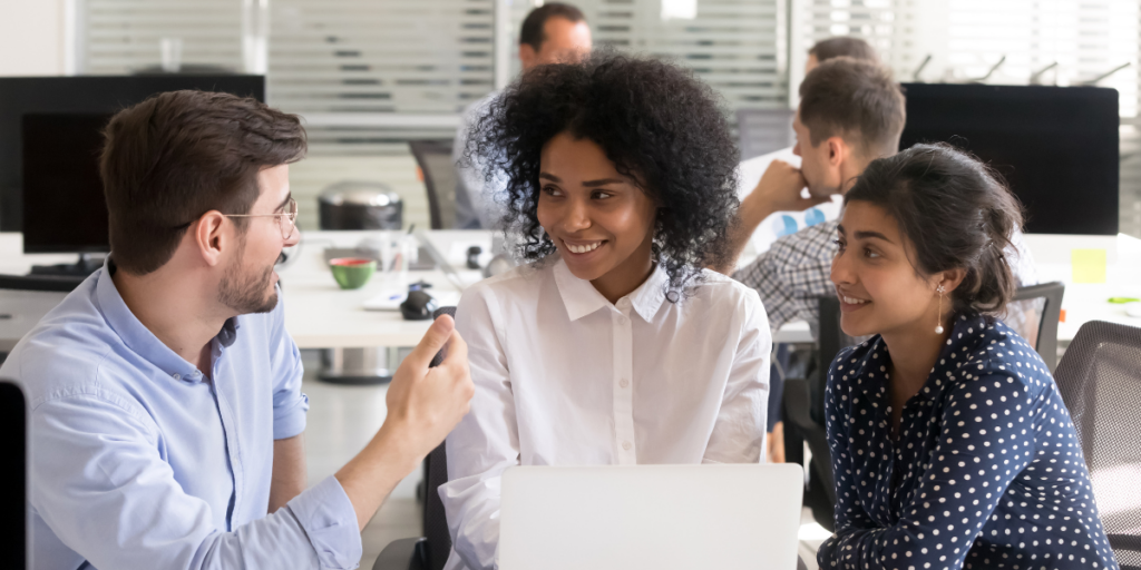 Three people are sat at a desk conversing and reviewing something on the laptop on the desk.