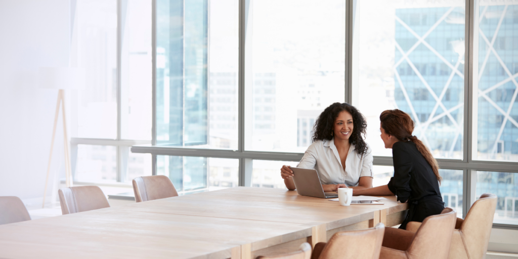 Two women sat in a boardroom in front of a wall of windows looking at a laptop.