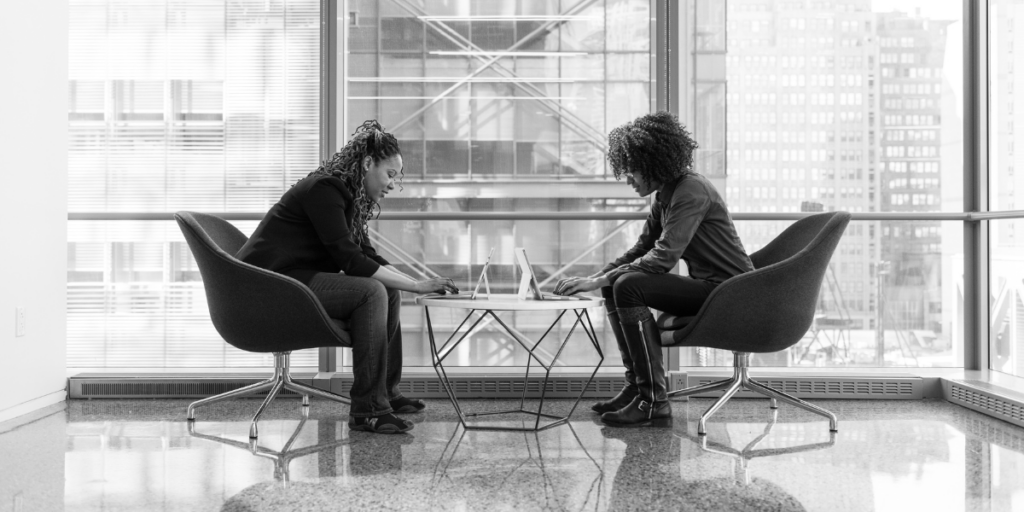 Two women are sat across from one another typing on their laptops. They are sitting in front of a wall of windows and the image is greyscale.