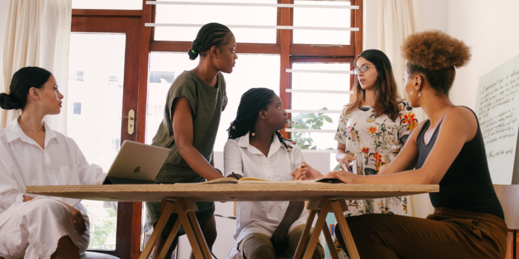 A group of women sitting at a boardroom table while one woman stands at a chart presenting.