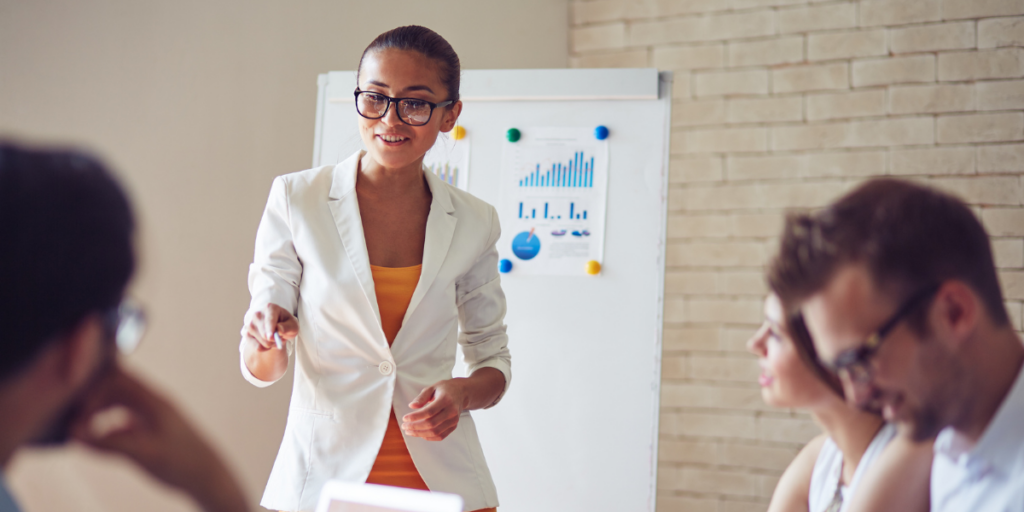 A woman standing in front of a chart at the front of a boardroom table presenting.