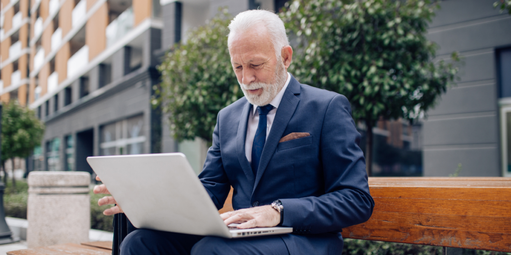 An older man sat on a bench typing on a laptop outside.
