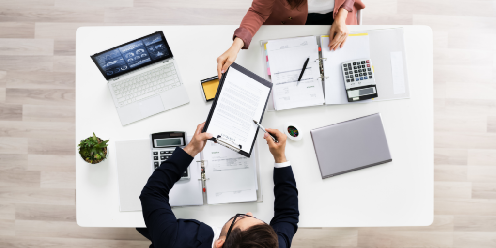 Two people are sat at a desk with paperwork and laptops. The one individual is showing the other a document on a clipboard.