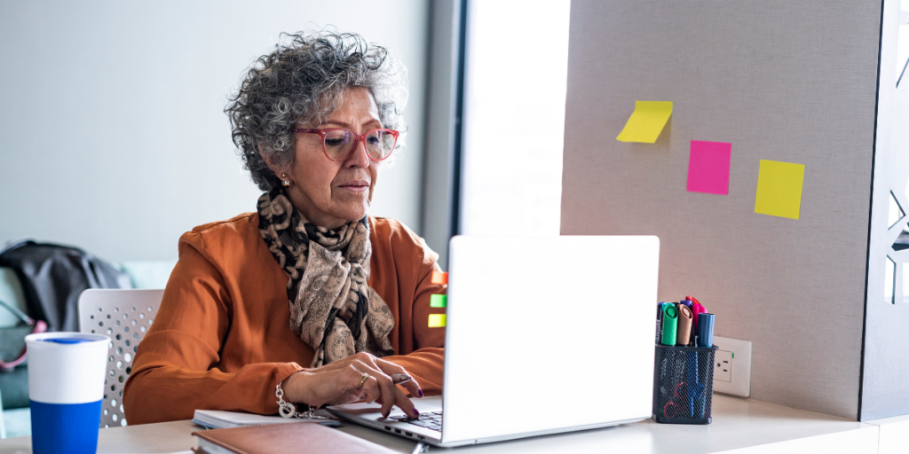 An older woman is sat typing on her laptop in her office.