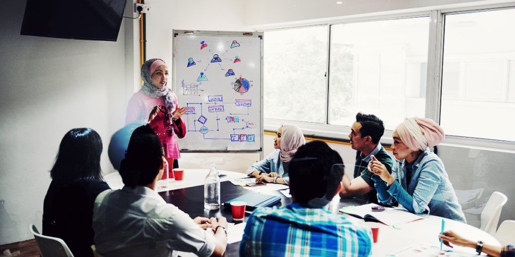A group of people sitting at a boardroom table. A woman is standing at a chart presenting.