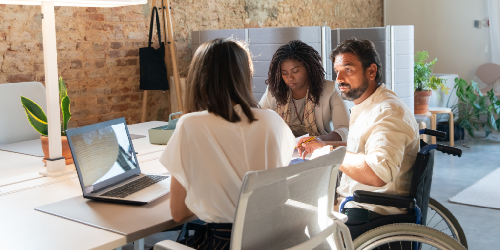 Two women are sat at a desk working, and one man in a wheelchair is talking with one of the women.