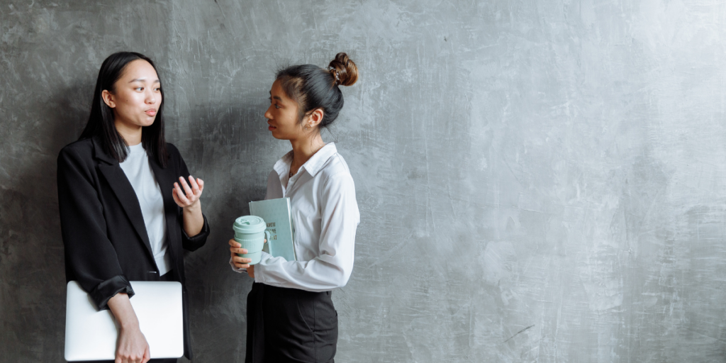 Two women are stood side-by-side along a grey wall conversing.