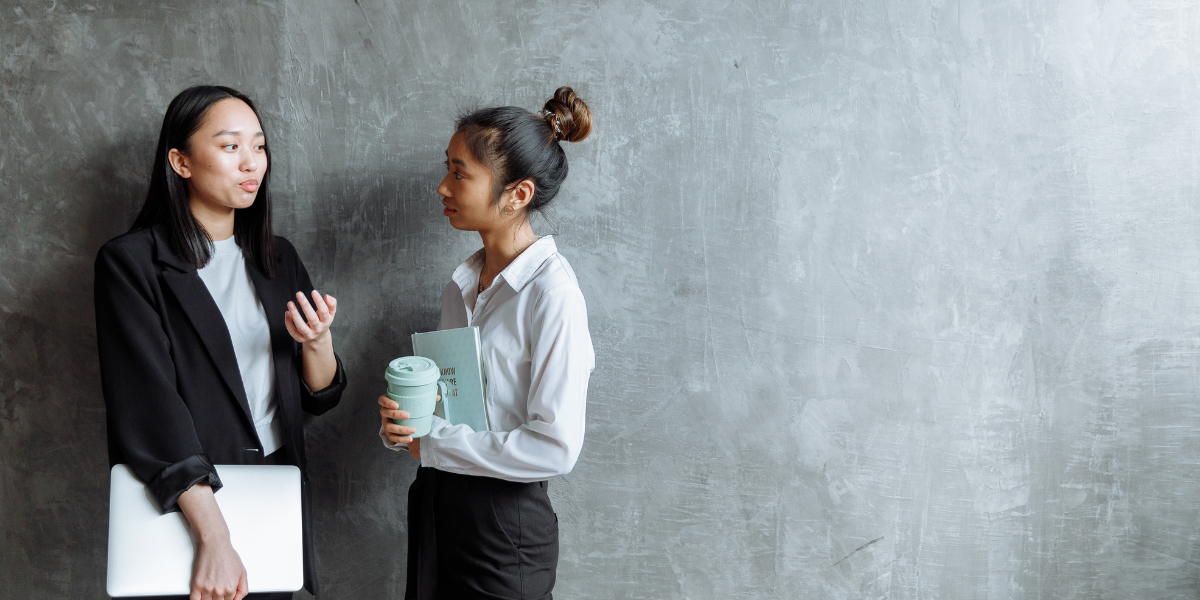 Two women are stood side-by-side along a grey wall conversing.