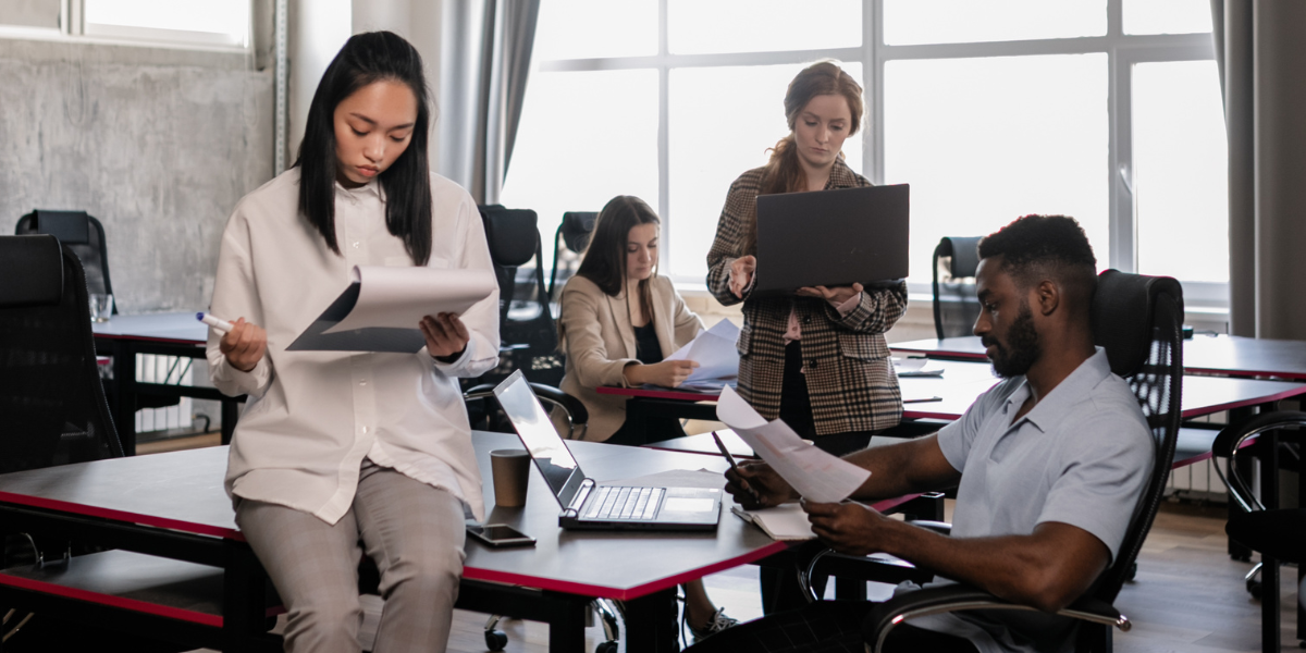 A group of people in an office space. A woman is sitting on the desk reviewing notes, and the others are working on their laptops.