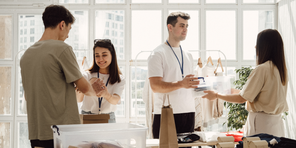 A group of four volunteers appear to be working a clothing drive. They are all standing working in front of a wall of windows, and they are handing boxes to one another.