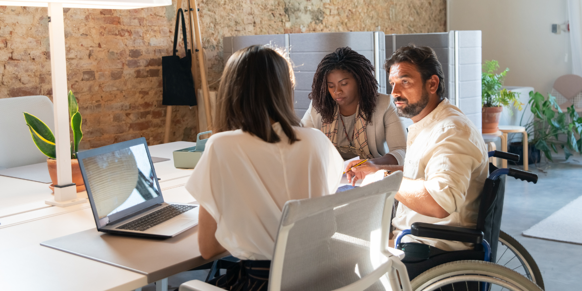 Two women are sat at a desk working, and one man in a wheelchair is talking with one of the women.