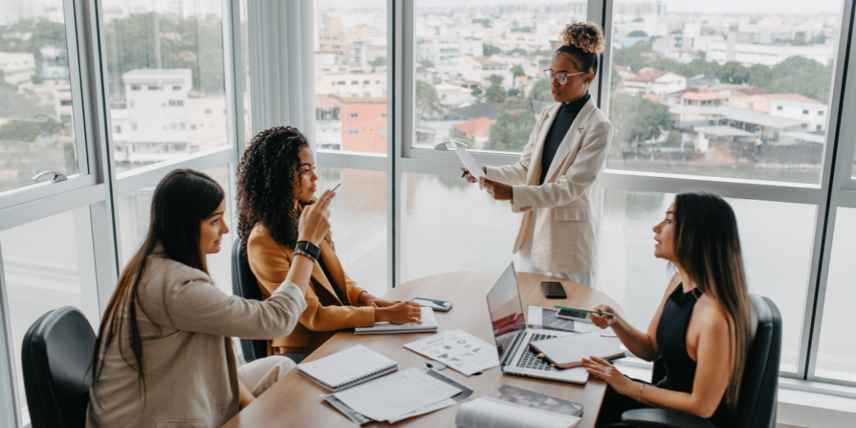 Three women sitting at a boardroom table and one woman is standing presenting to the group.