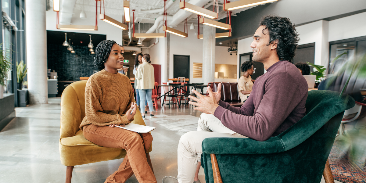 A man and a woman are sat in yellow and green velvet chairs - they are conversing.