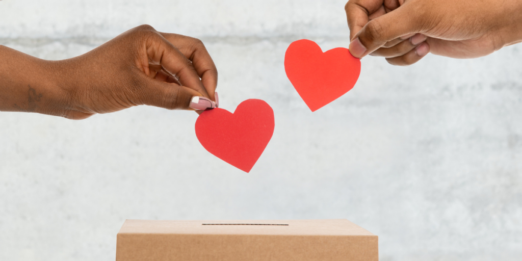 Two hands are holding red paper hearts and placing them in a brown ballot box.