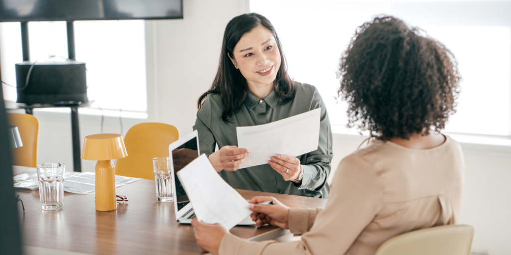 Two women are sat at a boardroom table. They are both holding pieces of paper and conversing.