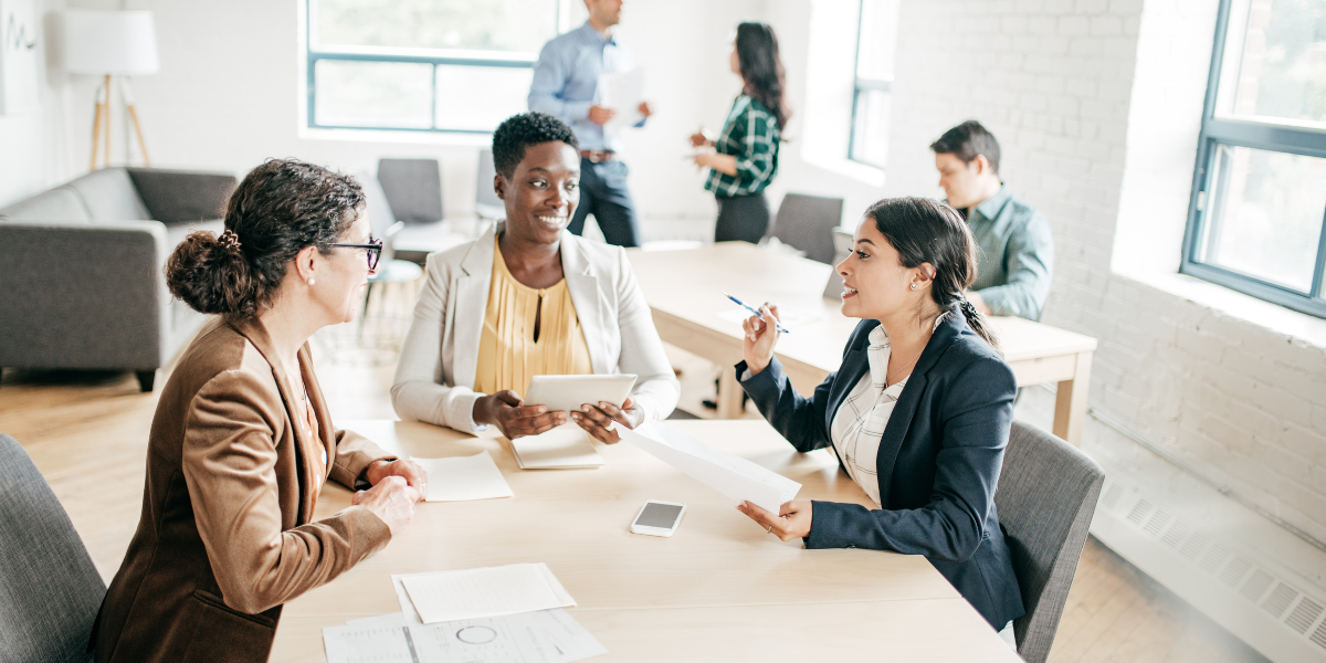 Three women are sat at a boardroom table. The one woman is holding a piece of paper and speaking.