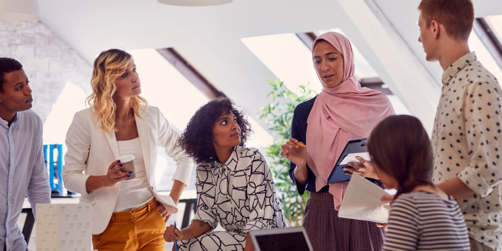 A large group of individuals are surrounding a boardroom table in a white dimly lit room. One of the women is stood holding a tablet and presenting to the team.