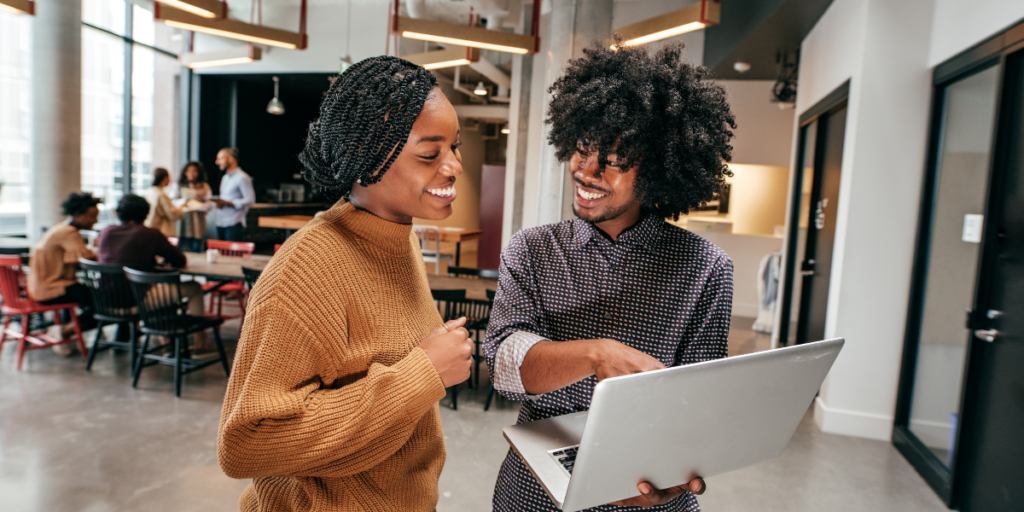 Two people are stood in an office. The man is holding a laptop and pointing at the screen while the woman looks on.
